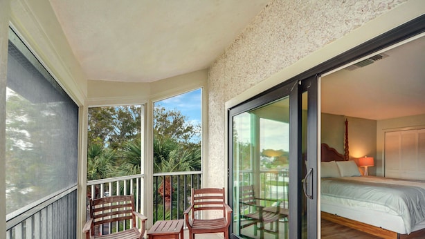 A balcony with wooden furniture, a glass door leading to a bedroom with a bed and a nightstand, and a view of palm trees. - On the Sea Palms Golf Course. 18th Hole, Fairway and Lagoon frontage (Saint Simons Island)