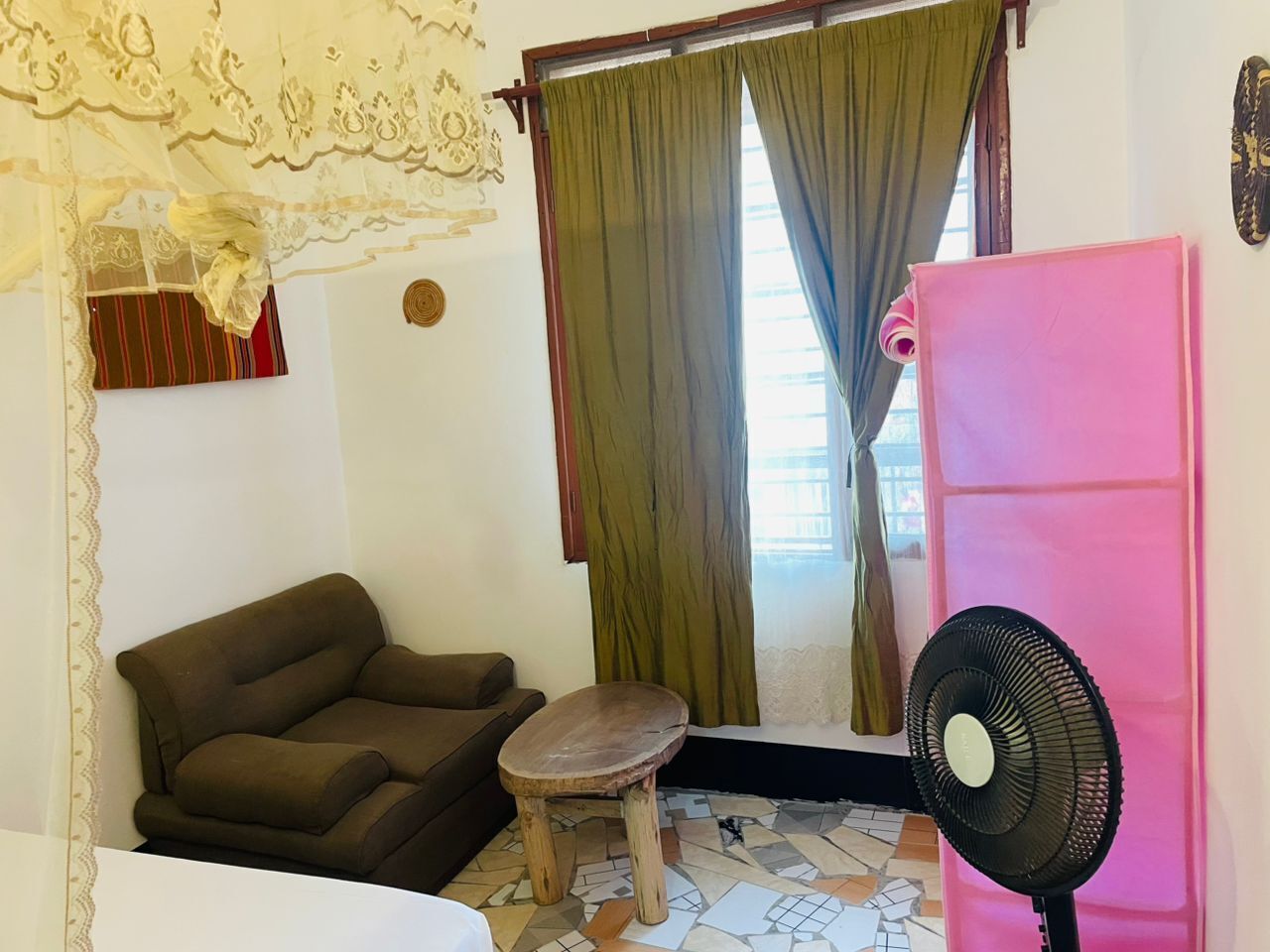 A room with a brown sofa, a small wooden stool, a fan, and a window with green curtains.