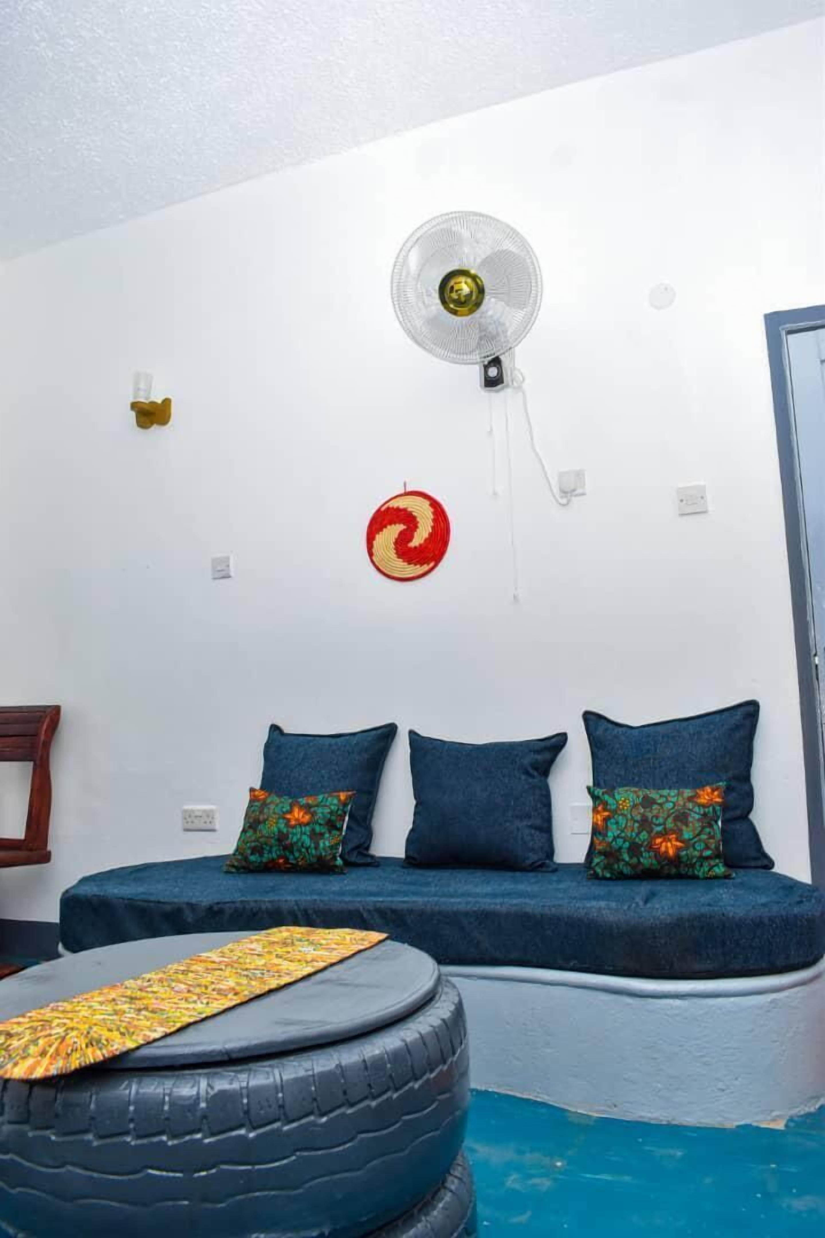 Una habitación con una cama, un ventilador, un tapiz en la pared y un otomán hecho con neumático.