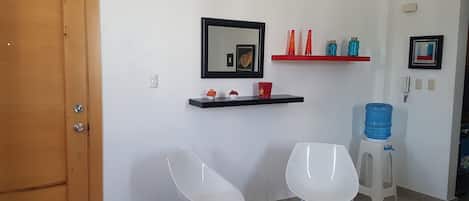 A clean, modern room with a wooden door, white walls, a black-framed mirror, a red shelf with decorative items, two white chairs, and a water dispenser.
