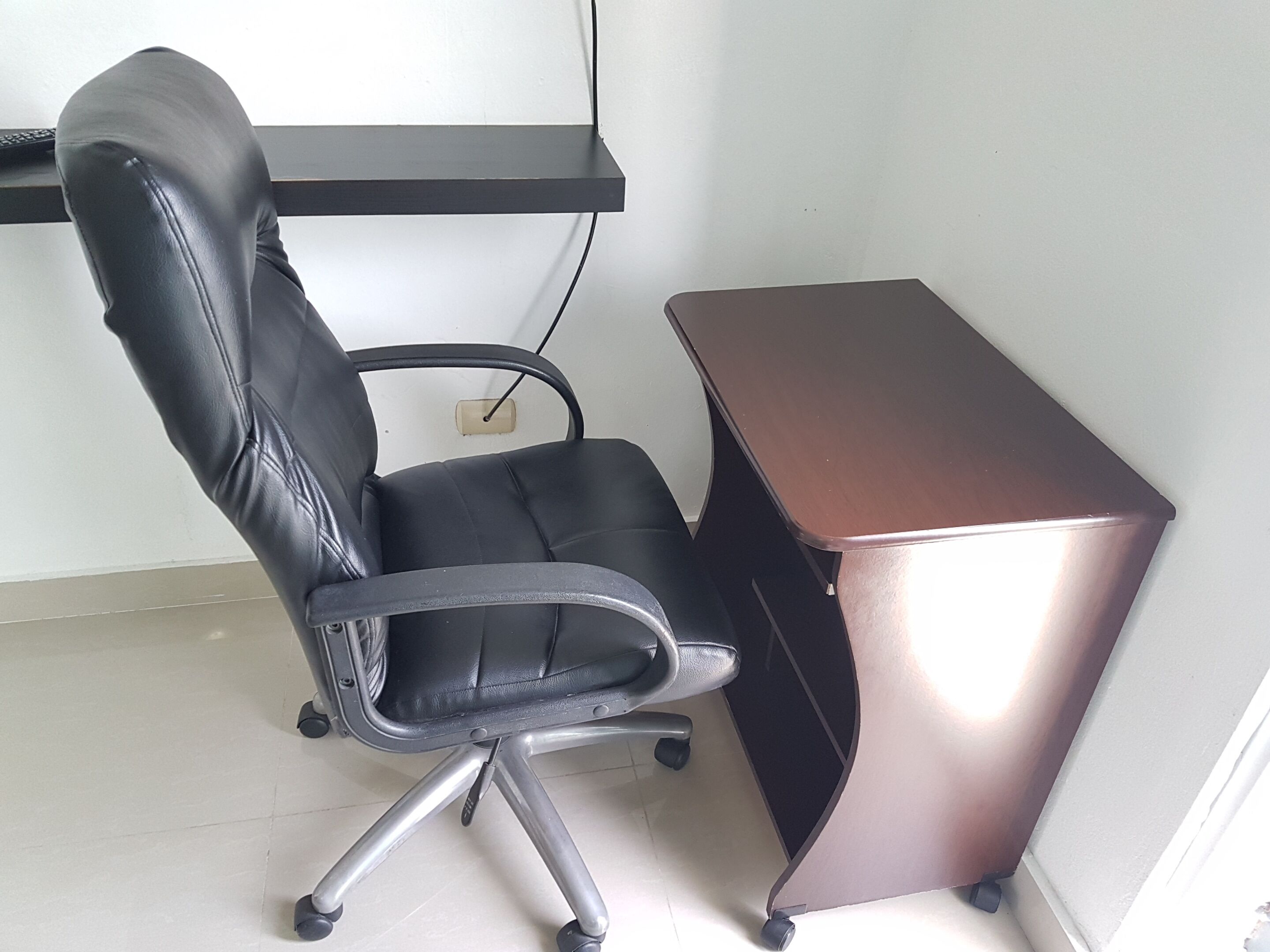 A black office chair with a five-point base, a brown desk, and a white wall with a power outlet.