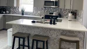 A modern kitchen with a large island, white cabinets, a microwave, and a sink. There are three stools around the island. - Cozy chic pool house 4 bed 2 bath KING BED in master bedroom (Bradenton)