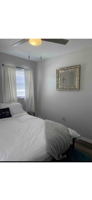 A bedroom with a bed, a ceiling fan, a window with curtains, and a mirror on the wall. - Cozy chic pool house 4 bed 2 bath KING BED in master bedroom (Bradenton)