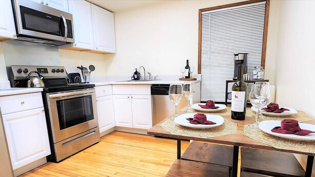 Una cocina moderna con electrodomésticos de acero inoxidable, armarios blancos y un comedor para dos personas, con platos y copas de vino.