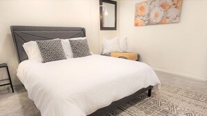 Una cama bien hecha con cabecera gris, ropa blanca y almohadas estampadas. Se ve una mesita de noche con una lámpara a la izquierda.
