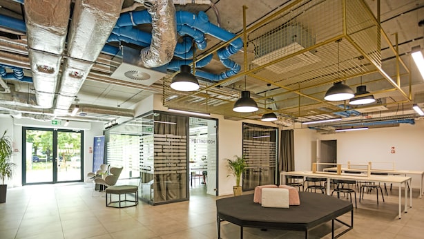 A modern office space with a meeting room, open tables, and a central seating area.