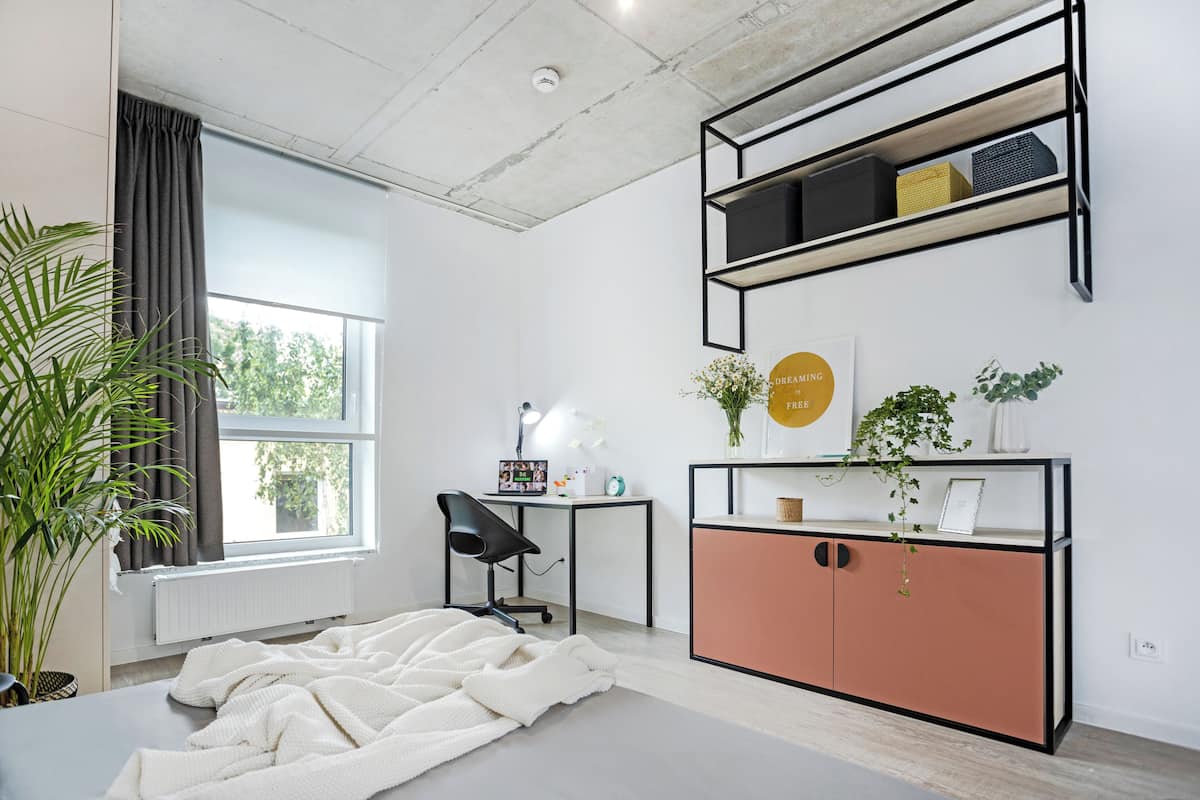 Habitación moderna con un ventanal grande, un escritorio con silla, un radiador blanco, una planta en maceta y un armario rosa con estantes.