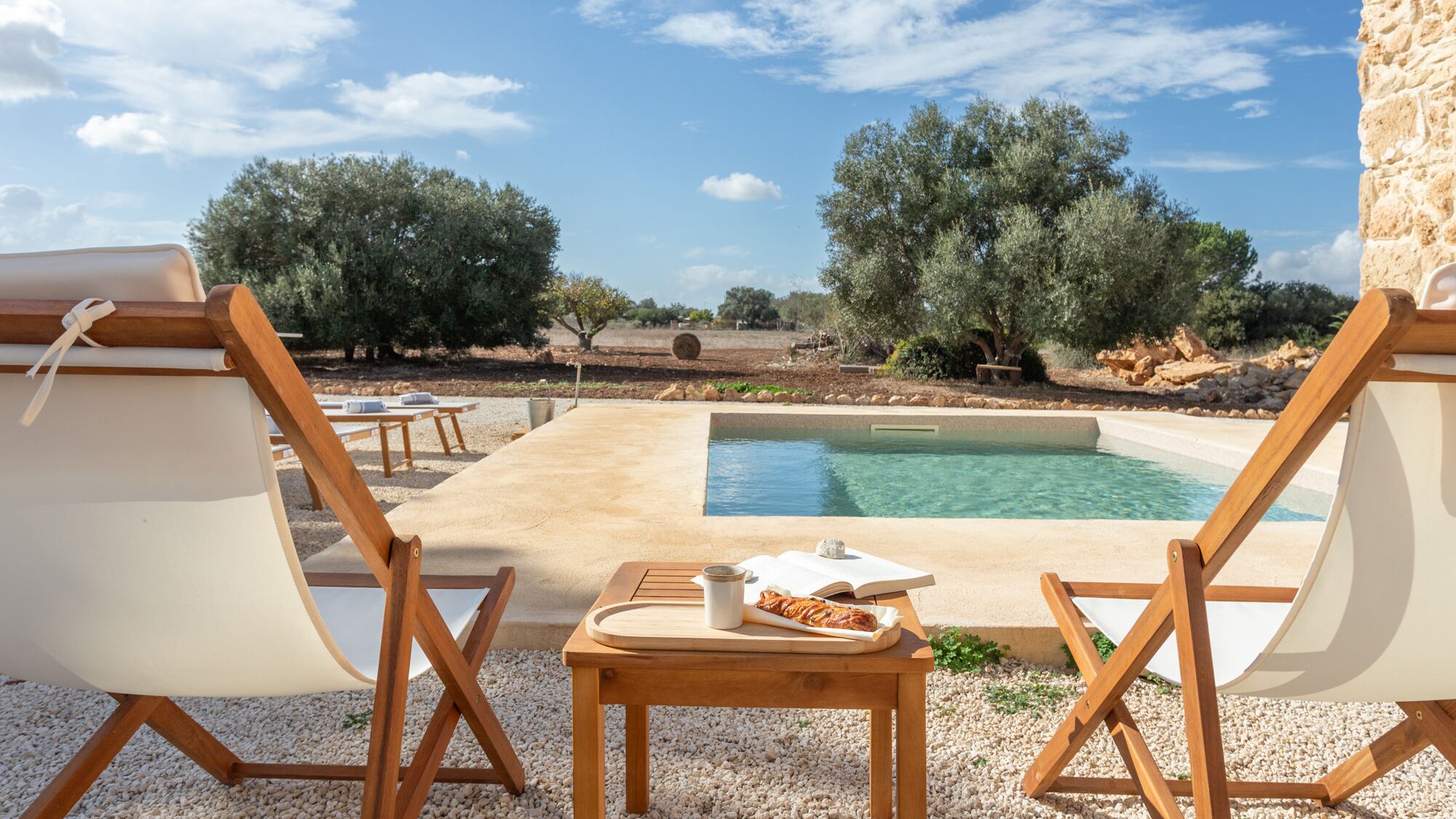 A poolside area with two lounge chairs, a small table with a tray of food, and a view of a pool surrounded by olive trees.