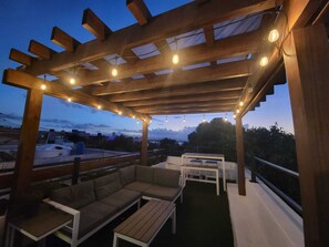 Een terras buiten met een houten pergola, lichtslingers, een bank en een tafel.