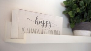 A white shelf with a sign that reads "happy is always a good idea" and a potted plant.