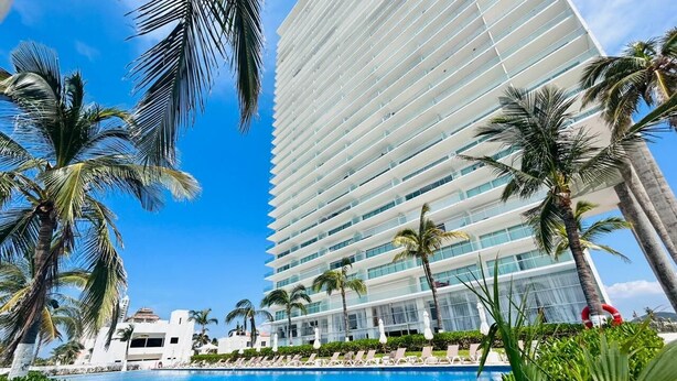 Un hotel moderno con una piscina grande, palmeras y un cielo azul despejado.