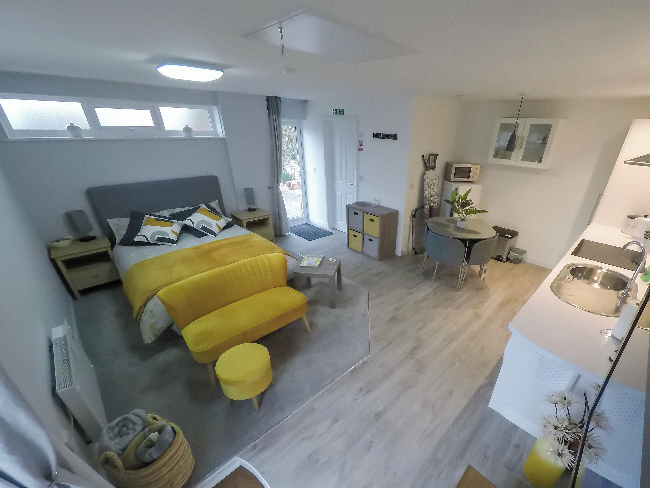 Ein modernes Hotelzimmer mit Bett, Nachttisch und kleinem Schreibtisch mit Stuhl. Ein Fenster bietet Blick ins Freie, es gibt eine Küchenzeile mit Spüle und einen kleinen Essbereich.