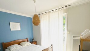A bedroom with a wooden bed, a bedside table, a hanging light, and a window with curtains. - Apartment with garden - sea view (Palavas-les-Flots)