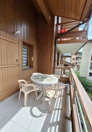 A wooden balcony with a round table and chairs, a window with a curtain, and a view of other buildings.