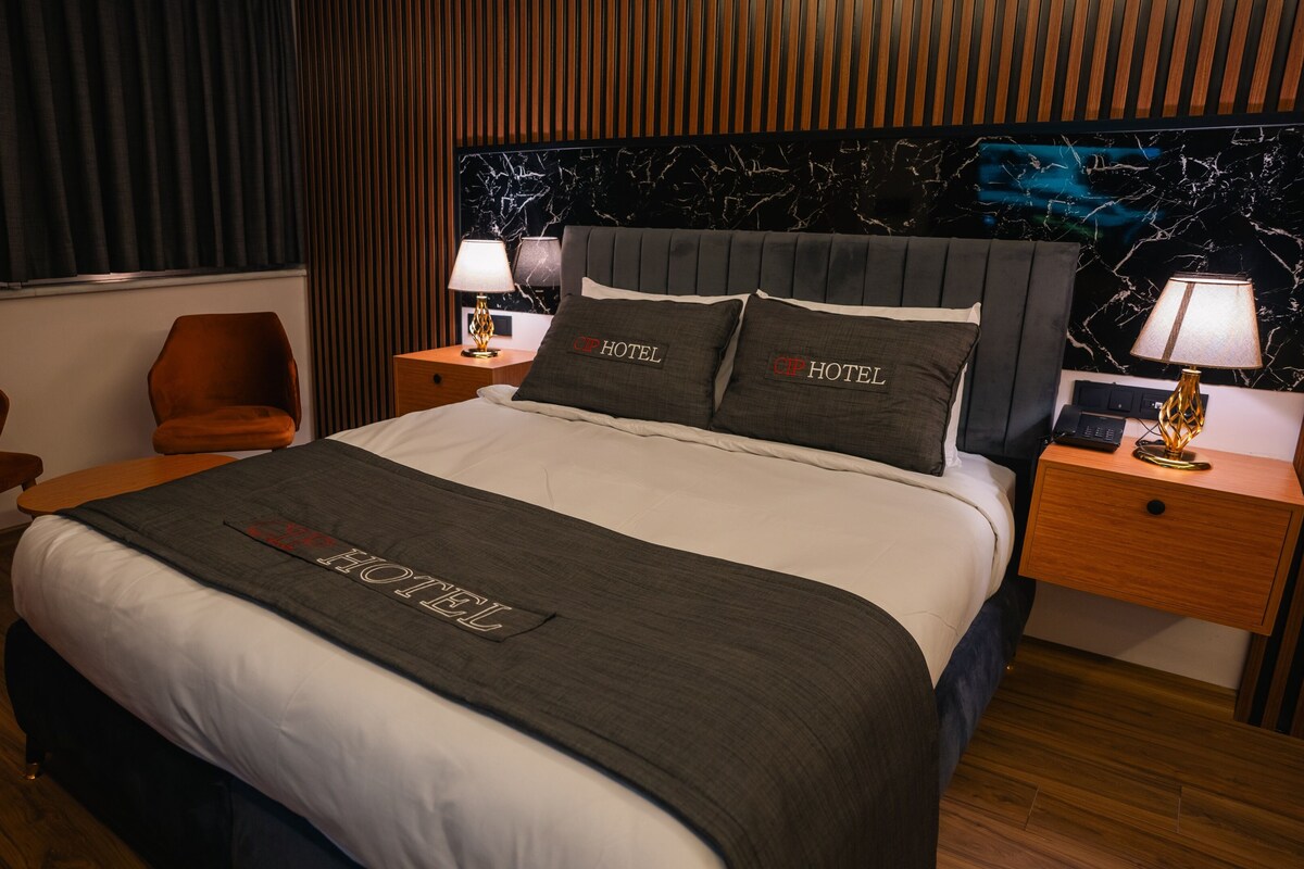 A hotel room with a bed, two chairs, a nightstand with a lamp, and a marble headboard.