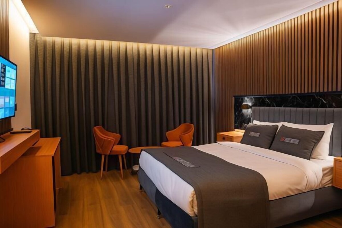 A hotel room with a bed, a desk, two chairs, a television, and a large window with curtains.