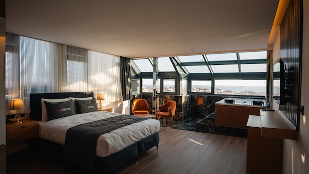 A modern hotel room with a large bed, two chairs, a desk, and a view of the city through large windows. - Cip hotel istanbul  (Istanbul)