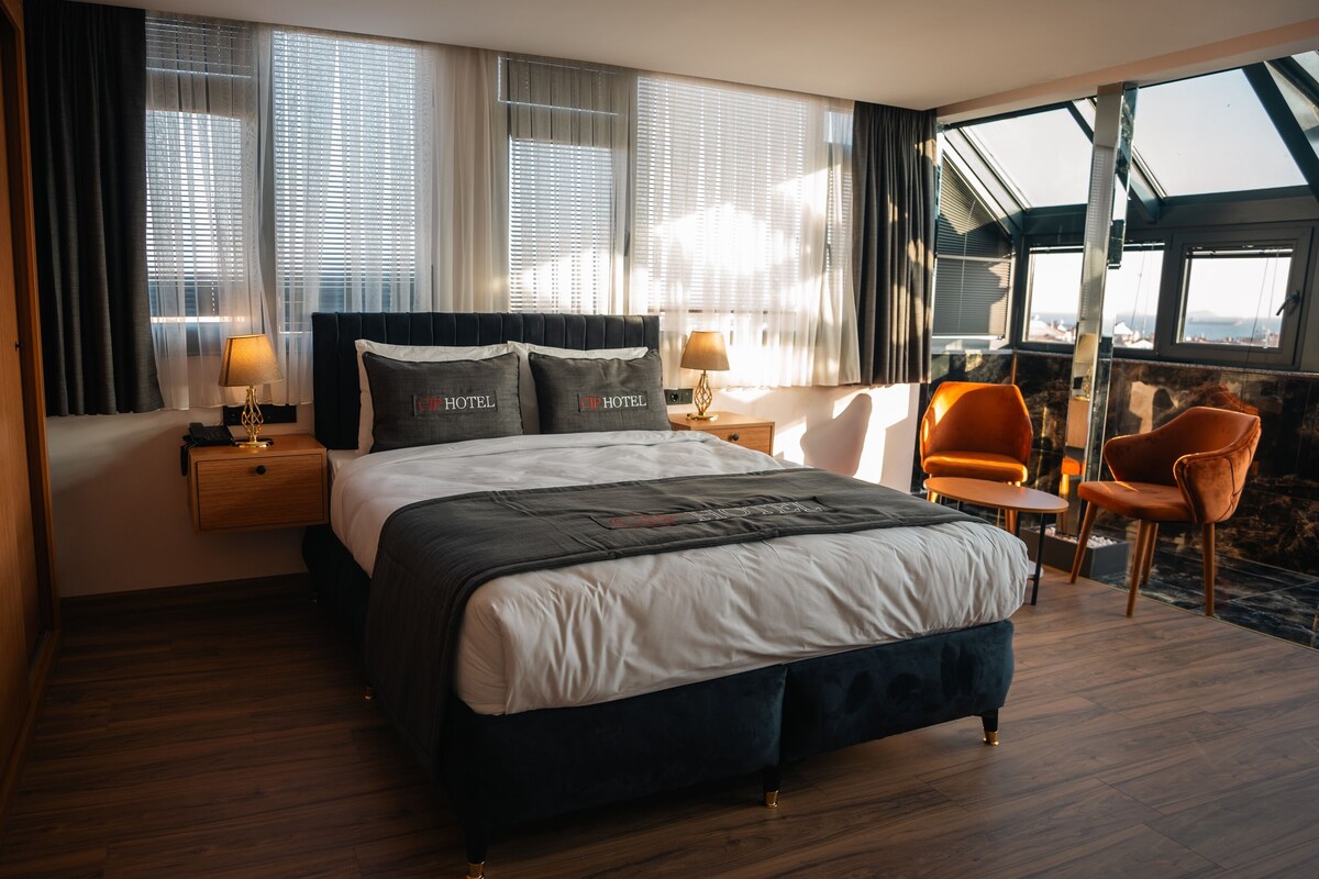 A modern hotel room with a large bed, bedside tables, a chair, and a view of the outdoors.