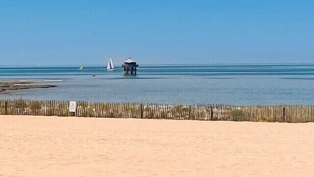 A beach with a wooden fence, a small structure, and a sailboat in the water. - Charming love nest with balcony (La Rochelle)