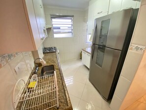 A kitchen with white cabinets, a stainless steel refrigerator, a gas stove, and a window with blinds. - Foot in the sand Corner of Fort II - Tiradentes 3 (Praia Grande)