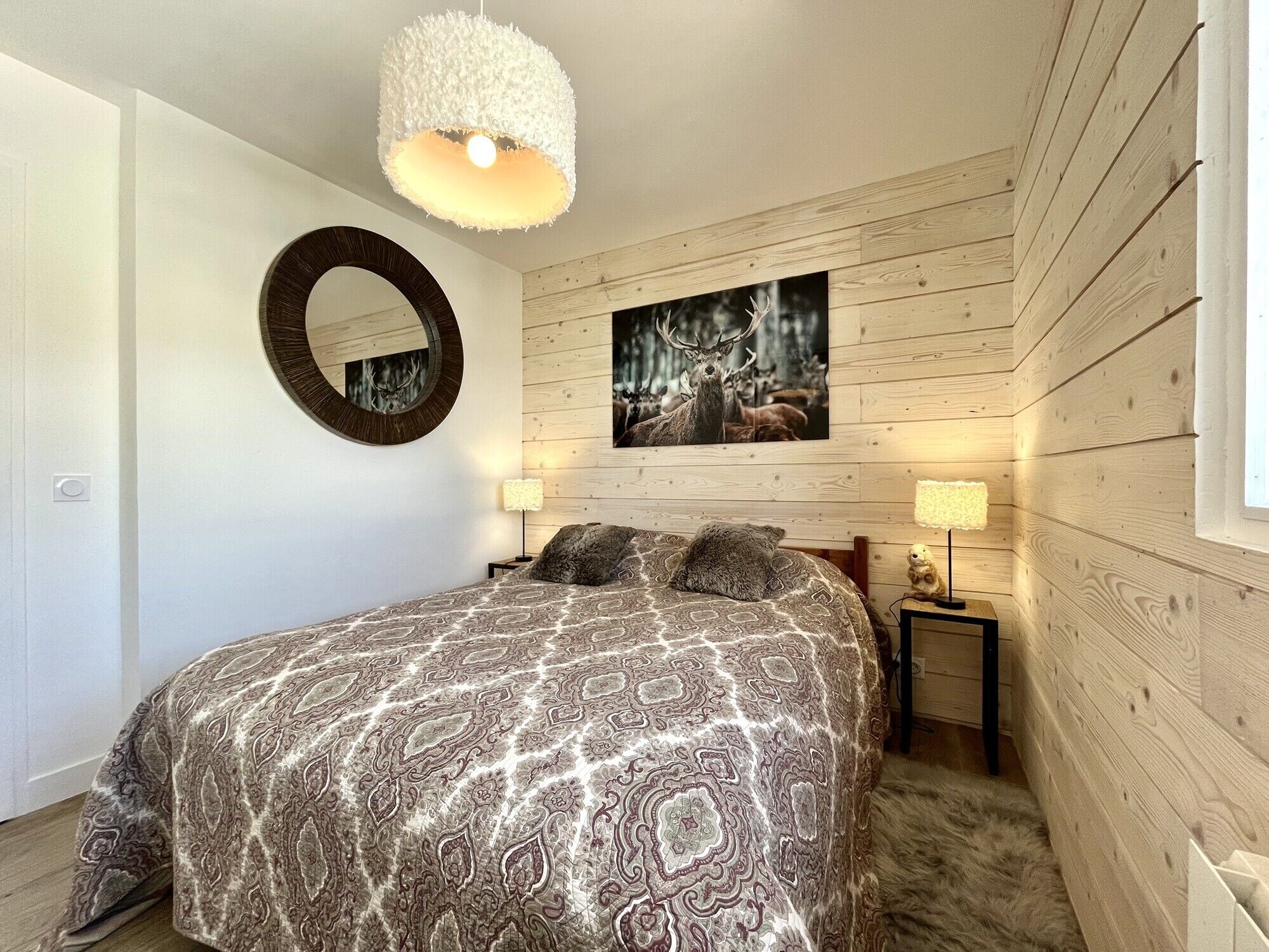 Un dormitorio con una colcha estampada, un espejo redondo, una fotografía enmarcada de fauna silvestre y paneles de madera en la pared.