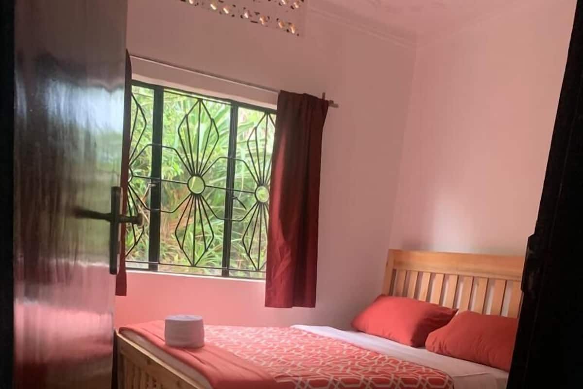 A bedroom with a wooden bed, red and white bedding, a window with a metal grill, and a pair of slippers on the floor.
