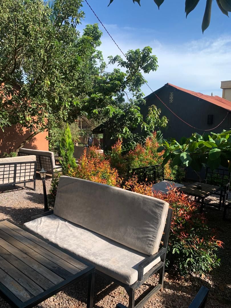 A wooden bench with a cushion in a garden area with various plants and a building in the background.