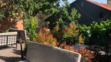 A wooden bench with a cushion in a garden area with various plants and a building in the background.