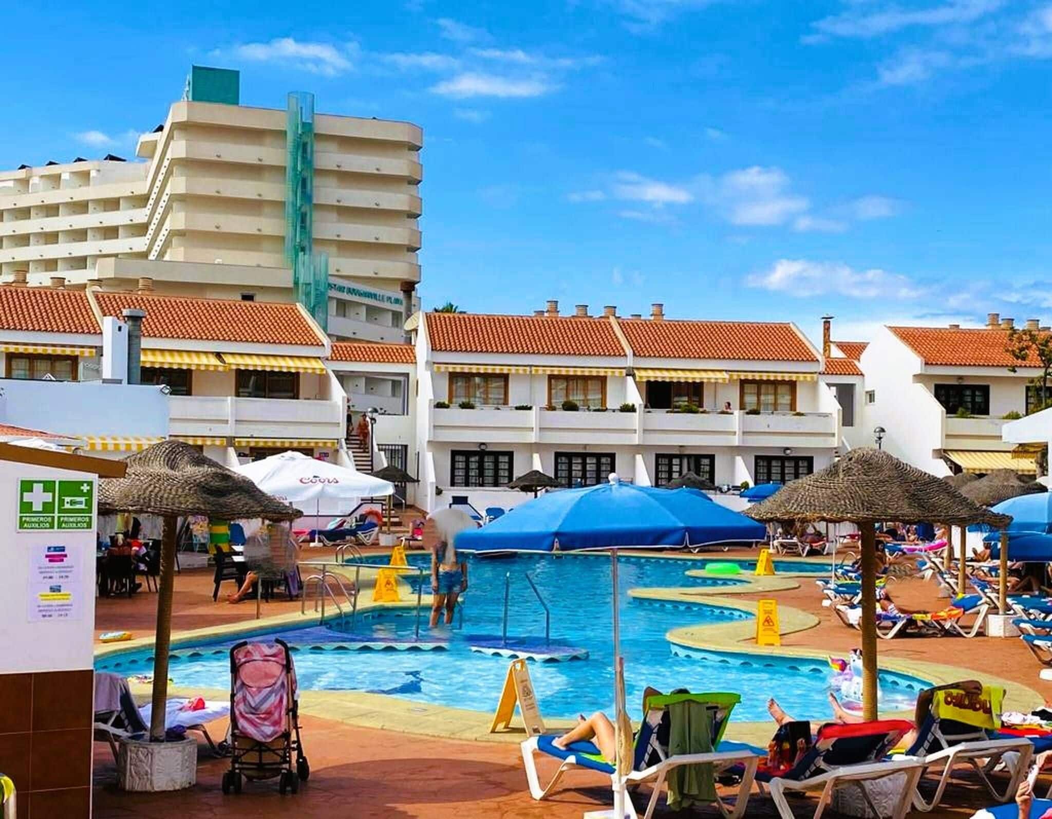 A hotel pool area with multiple swimming pools, sun loungers, umbrellas, and a building with a tiled roof.