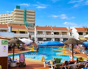 Zona de piscina de hotel con varias piscinas, tumbonas, sombrillas y un edificio con tejado de azulejos.