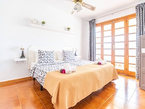 Quarto com cama, ventilador de teto, janela com cortinas e uma pequena prateleira com objetos decorativos.