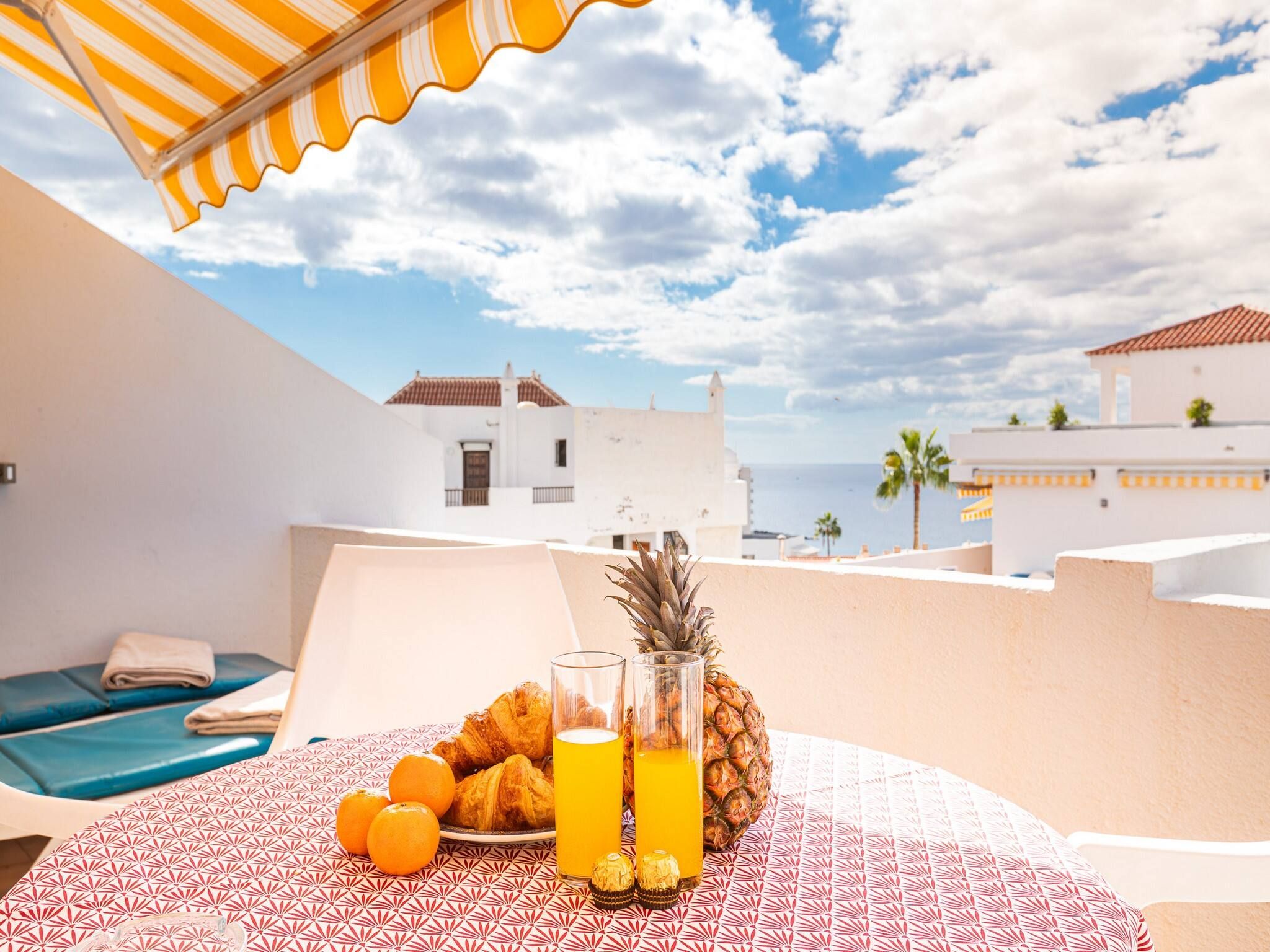 A table with breakfast items including fruits, a pineapple, and orange juice, set against a backdrop of a white building and a clear sky.