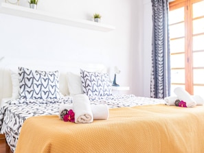 Habitación de hotel con cama, almohadas, mesita de noche, planta en maceta y ventana con cortinas.