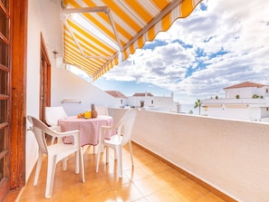Uma varanda com mesa e cadeiras, um toldo listrado e vista para edifícios sob um céu parcialmente nublado.