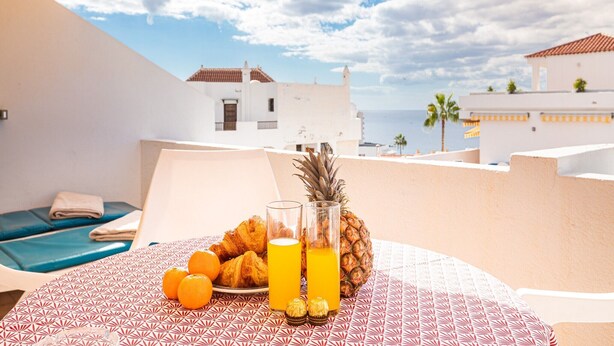 Ein Tisch mit Croissants, Orangen, einer Ananas und einem Glas Orangensaft, bedeckt mit einer gemusterten Tischdecke, mit Blick auf eine Küstenstadt mit weißen Häusern und blauem Himmel.