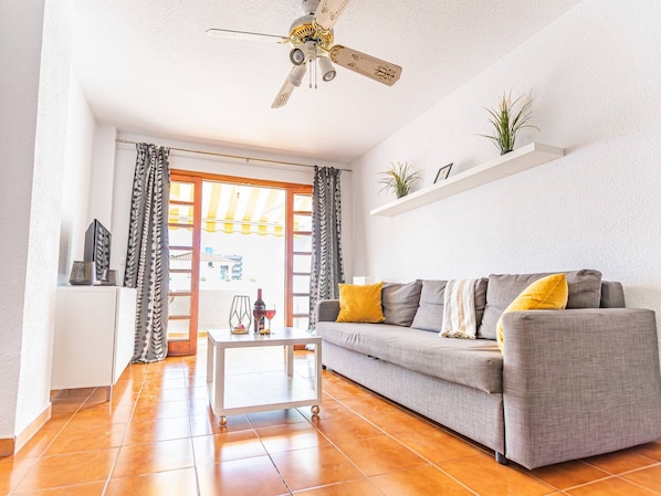 Una sala de estar con un sofá gris, una mesa de centro blanca, un ventilador de techo y un ventanal con cortinas.