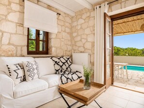 A room with a stone wall, a white sofa with patterned cushions, a wooden coffee table, a radiator, and a view of a swimming pool.