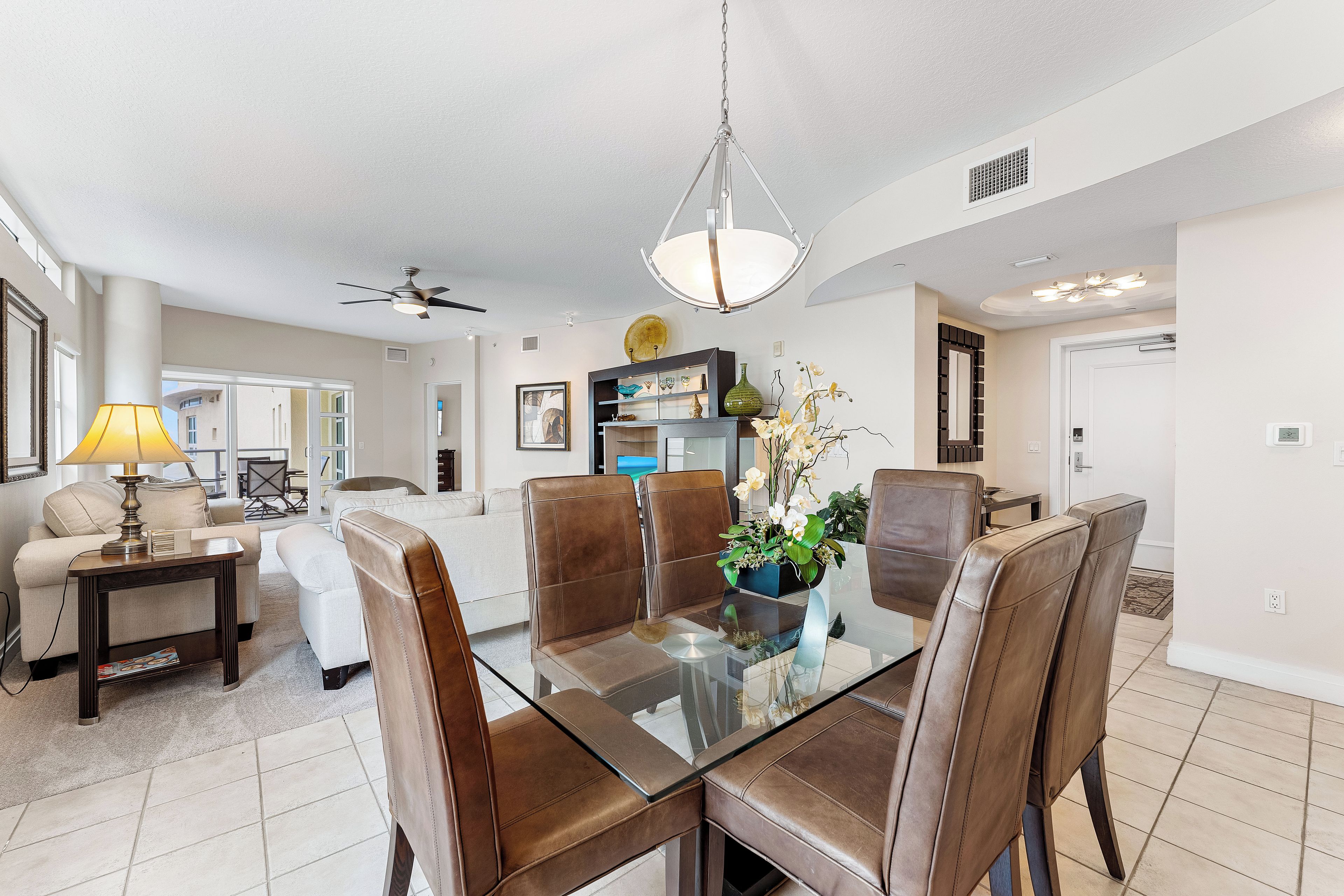 A modern dining area with a glass table and leather chairs, a ceiling fan, and a decorative plant centerpiece.