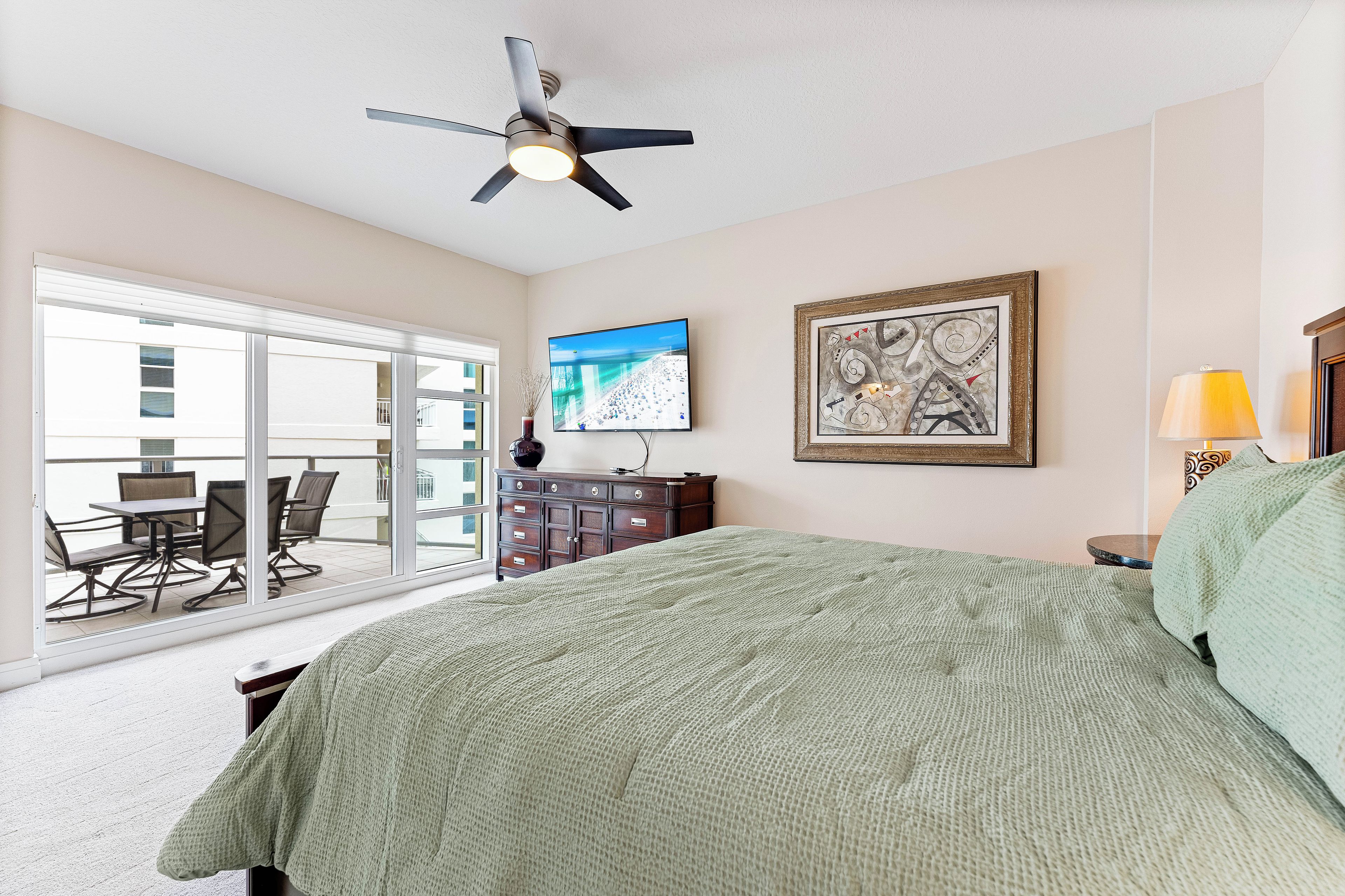 A bedroom with a large bed, a ceiling fan, a flat-screen TV, a dresser, and a balcony with outdoor seating.