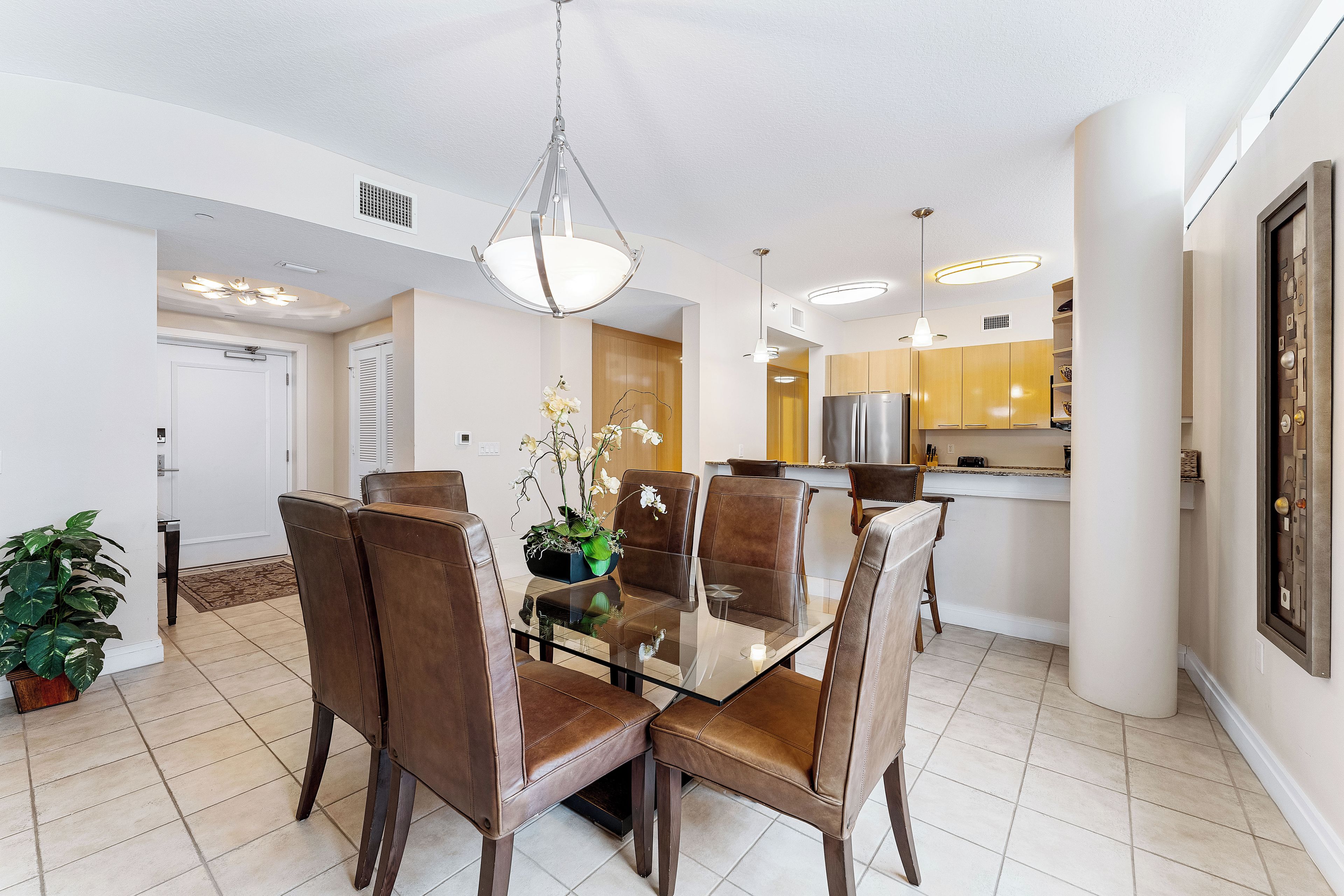 A modern dining area with a glass table and leather chairs, a hanging light fixture, and a kitchen in the background.