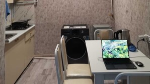 Cocina compacta con estufa a gas, microondas y refrigerador pequeño. Hay una mesa de comedor con una laptop y un ratón, y una silla. La habitación tiene un papel tapiz con textura y una lámpara de techo.