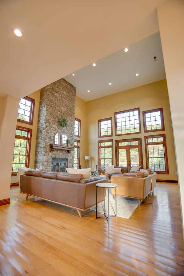 A spacious living room with a stone fireplace, large windows, and a high ceiling.