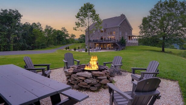 A stone fire pit surrounded by Adirondack chairs, a wooden table, and a large stone building in the background.