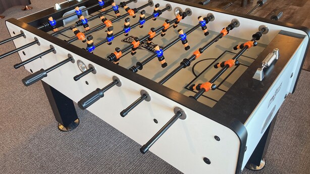 Une table de baby-foot dotée de plusieurs tiges et de plusieurs figurines, posée sur un sol recouvert de moquette.