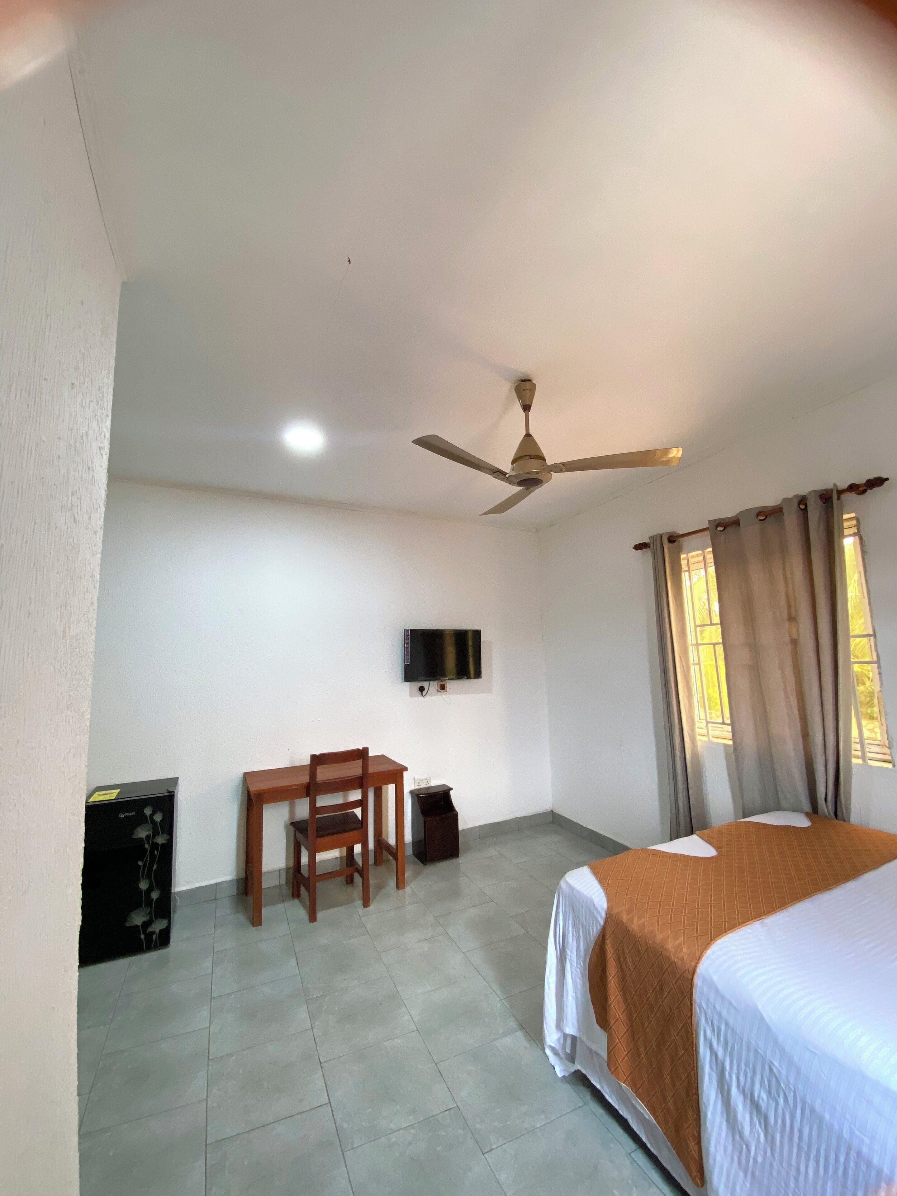 A hotel room with a bed, a desk with a chair, a ceiling fan, a TV mounted on the wall, and a window with curtains.