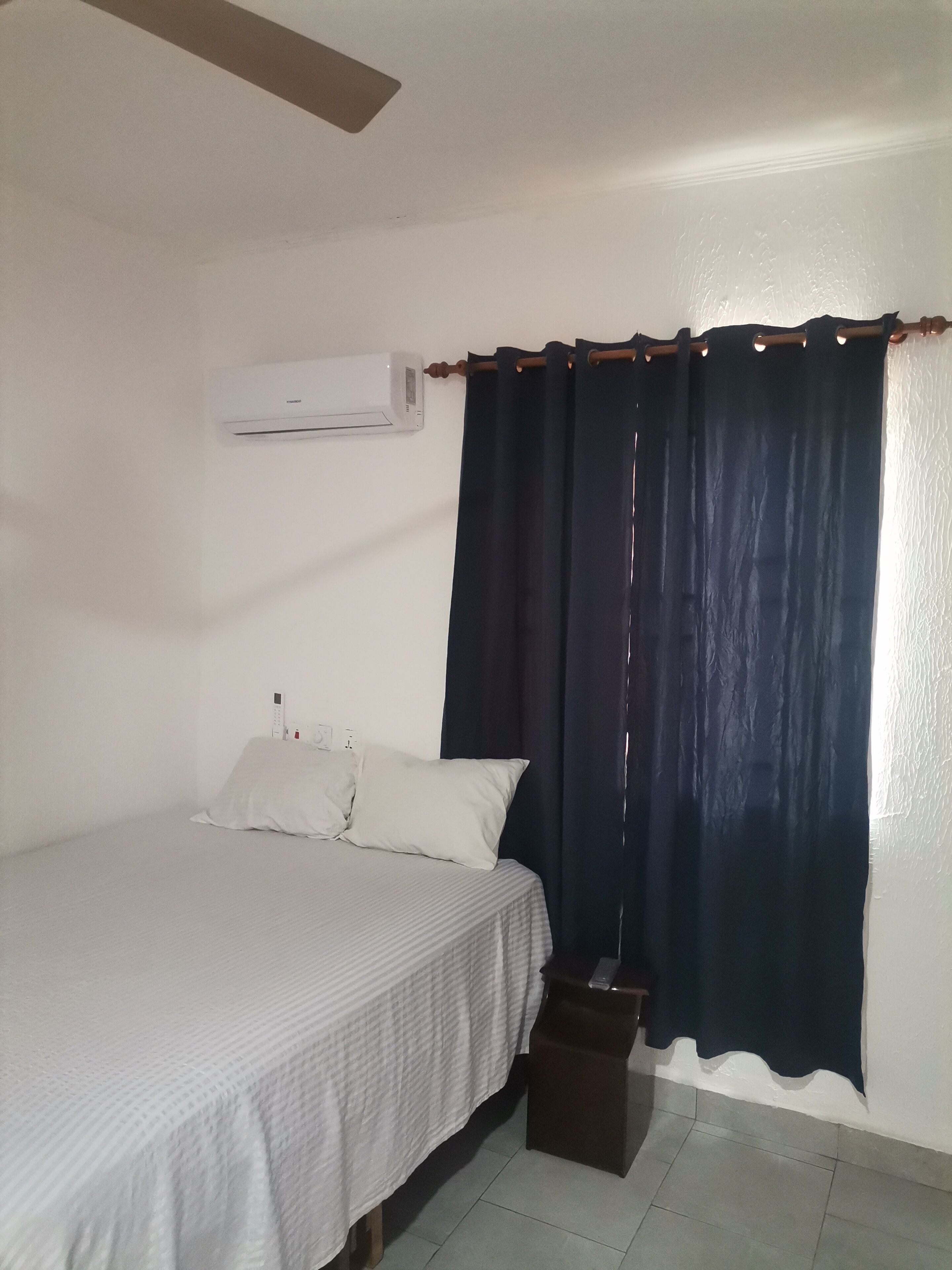 Quarto com cama, roupa de cama branca, cortina escura, um banquinho de madeira pequeno e um aparelho de ar condicionado na parede.