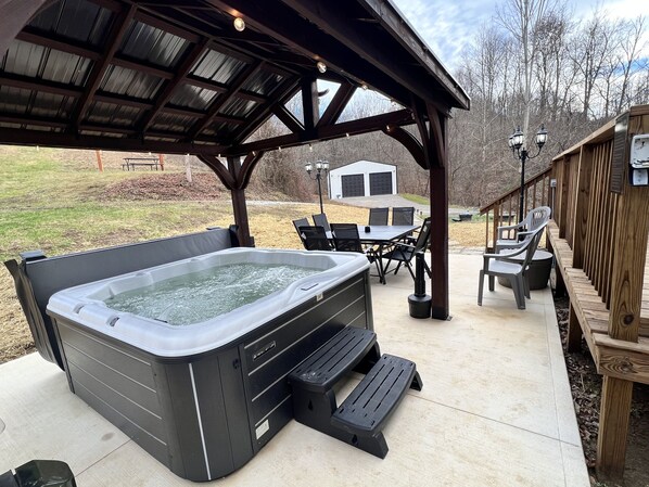 A covered outdoor hot tub area with a table and chairs, a wooden deck, and a garage in the background.