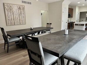 Un espace repas avec une longue table et des chaises, une cuisine à l’arrière-plan et une œuvre d’art accrochée au mur.