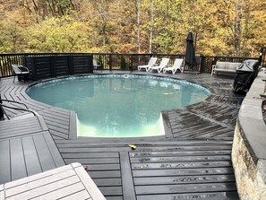 A circular pool with a wooden deck, surrounded by white lounge chairs and a covered seating area.
