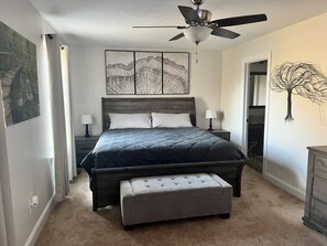 Ein Schlafzimmer mit einem Bett, einem Deckenventilator, einem Nachttisch, einer Bank und einer Wand mit einem Baumgemälde.